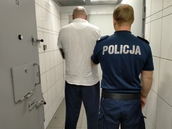 Na zdjęciu widać umundurowanego policjanta z zatrzymanym w korytarzu policyjnego aresztu, obaj stoją tyłem.