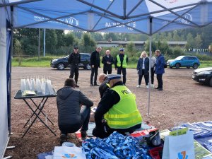 policjant i strażak klęczą przy fantomie, tłumaczą mężczyźnie zasady udzielania pierwszej pomocy, policjanci i inne osoby stoją niedaleko nich i obserwują