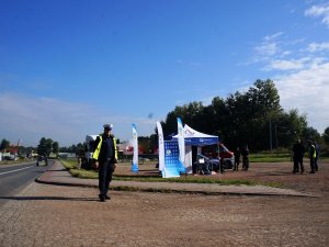 policjant w mundurze stoi przy drodze w tle namiot policyjny, strażak i mężczyzna kucają, niedaleko nich stoją inni policjanci, strażacy i osoby i ich obserwują