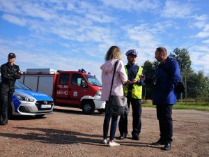 policjant rozmawia z kobietą i mężczyzną, w tle strażak, i radiowóz policyjny oraz wóz strażacki