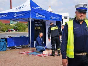 policjant w mundurze uśmiecha się, w tle mężczyzna klęczy przed fantomem położonym na kocu, pod namiotem z napisami policja