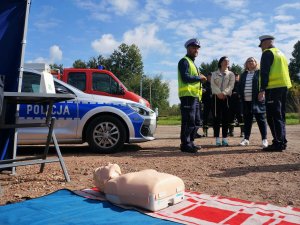 na kocu położony fantom, policjanci w mundurach rozmawiają z kobietami, obok stoi radiowóz policyjny