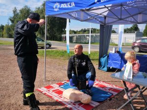 mężczyzna klęczy na kocu przed fantomem, przed nim stoi strażak, obok stoi mała dziewczynka, nad nimi namiot z napisem policja