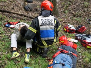 strażak rozmawia z osoba poszkodowaną, która trzyma się za głowę, obok leży manekin na desce ortopedycznej