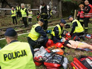 umundurowani policjanci klęczą przy osobach poszkodowanych, strażak idzie po kolejnych poszkodowanych, inne osoby w kamizelkach obserwują ćwiczenia