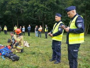 policjanci umundurowani stoją, jeden z nich rozmawia przez radiotelefon, stoi wóz straży pożarnej, pozoranci stoją w oddali, dwóch strażaków kuca, jeden pije wodę z butelki