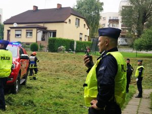 umundurowany policjant rozmawia przez radiotelefon, w tle widać pozostałych policjantów i wóz stażacki