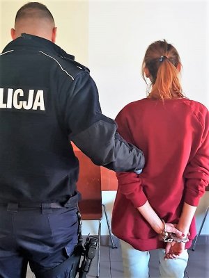 zatrzymana kobieta z kajdankami na nadgarstkach stoi tyłem, za ramie trzyma ją umundurowany policjant, też stoi tyłem