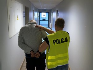 policjant w kamizelce odblaskowej z napisem Policja trzyma za ramię mężczyznę skutego w kajdanki