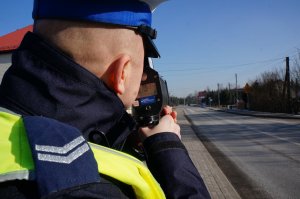 umundurowany Policjant mierzy prędkość pojazdów urządzeniem do pomiaru prędkości, stoi przy drodze