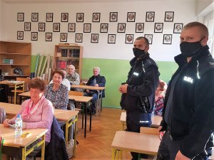 seniorzy na spotkaniu z dzielnicowymi siedzą w ławkach szkolnych, umundurowani Policjanci uśmiechają się do zdjęcia