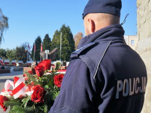 Policjant trzyma kwiaty w kolorach biało- czerowonych na tle flagi państwowej