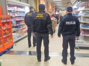 policjant i 2 strażników miejskich znajdują się w sklepie, stoją tyłem, sprawdzają przestrzeganie przez klientów obowiązku zakrywania ust i nosa przy pomocy maseczki