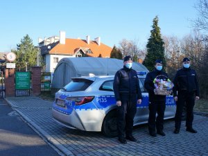 policjanci stoją przed radiowozem, policjant stojący w środku trzyma kosz ze słodkościami, w tle widać budynek domu pomocy społecznej