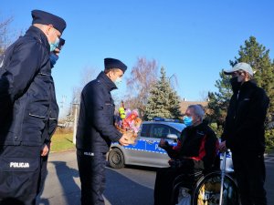 trzech policjantów stoją, z których jeden przekazuje kosz ze słodkościami seniorowi siedzącemu na wózku inwalidzkim, w tle widać budynek domu pomocy społecznej