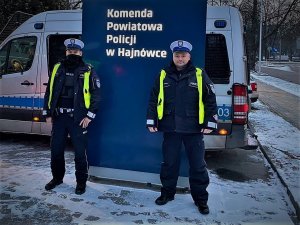 Zdjęcie przedstawia dwóch policjantów wydziału ruchu drogowego z Wodzisławia Śląskiego, którzy stoją przy tablicy Komendy Powiatowej Policji w Hajnówce