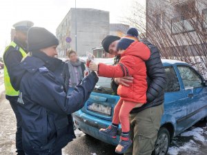 policjantka przekazuje odblaskową maskotkę dziecku, rodzice stoją obok zaparkowanego samochodu, widać też policjantów z drogówki, którzy stoją obok