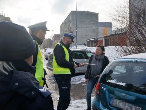 policjanci z drogówki rozmawiają z kierującą, która stoi obok swojego auta, policjant przekazuje pakiet z zawartością ulotki informacyjnej dot. kampanii informacyjno-edukacyjnej &quot;Nie pije. Prowadzę. Żyję&quot;