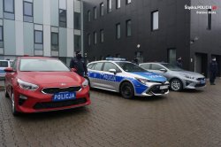 przed budynkiem Komendy Wojewódzkiej Policji w Katowicach stoją obok siebie trzy radiowozy, dwa nieoznakowane i jeden radiowóz oznakowany, obok radiowozu stoi umundurowany policjant