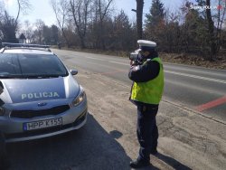 obok policyjnego radiowozu stoi policjant, w rękach trzyma urządzenie do pomiaru prędkości, stoi przy drodze, kontroluje prędkość pojazdów