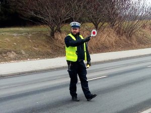 umundurowany policjant drogówki stoi na pasie ruchu i daje sygnał do zatrzymania się kierującemu pojazdem, podnosi tarczę do zatrzymywania pojazdów, w drugiej ręce ma alkomat