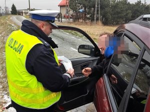 umundurowany policjant przekazuje pakiet z gadżetami kierowcy zatrzymanemu do kontroli drogowej, kierowca ma otwarte przednie drzwi samochodu, uśmiecha się do policjanta