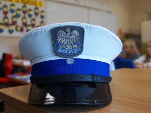 czapka policjanta drogówki na pierwszym planie, w tle klasa lekcyjna, szkolna gazetka i dzieci siedzące w ławkach