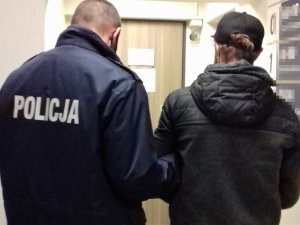 umundurowany policjant trzyma za ramię zatrzymanego mężczyznę, oboje stoją tyłem w korytarzu komedny