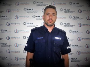 Dzielnicowy mł. asp. Wojciech Stencel na tle rollup z logo Komendy Powiatowej Policji w Wodzisławiu Śląskim