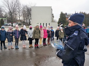 policjantka stoi na boisku szkolnym przed grupą dzieci, opowiada im o bezpieczeństwie podczas ferii zimowych