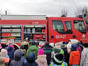 przed wozem strażackim stoi strażak, przed nim grupa dzieci słucha jego opowiadania