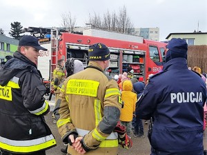 na zdjęciu widać, jak grupa dzieci stoi przed strażakiem, który im opowiada o bezpieczeństwie podczas ferii, na pierwszym planie stoją tyłem policjant i dwóch strażaków