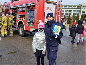 policjantka pozuje do zdjęcia z uczennicą, policjantka trzyma w ręku laurkę. W tle stoi wóz strażacki, strażacy i uczniowie.