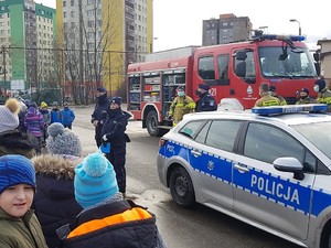 Policjantka i policjant stoją przed grupą dzieci, za nimi stoi policjant ze strażakami. Obok stoi policyjny radiowóz, w tle stoi wóz strażacki.