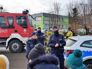 policjantka i policjant stoją przed grupą dzieci, rozmawiają z nimi, w tle widać wóz stażacki