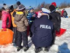 policjant w mundurze znajduje się na sztucznym stoku wraz z uczestnikami półkolonii, policjant kuca, rozmawia z dziećmi