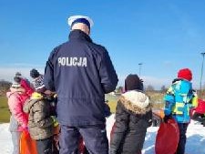 policjant w mundurze stoi tyłem, przed nim dzieci stoją i słuchają o czym opowiada