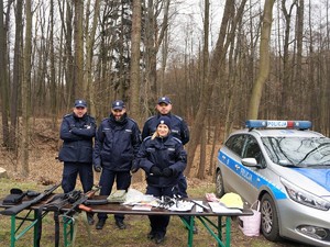 Troje policjantów i policjantka pozują do zdjęcia. Stoją obok policyjnego radiowozu, za stolikiem wypełnionym ulotkami i odblaskowymi gadżetami. Na stole znajdują się również jednostki broni.