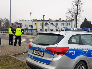 Na zdjęciu widać, jak w rejonie przejścia dla pieszych policjanci przyglądają się uczestnikom ruchu drogowego, niedaleko nich stoi zaparkowany radiowóz. Policjanci mają na sobie kamizelki odblaskowe z napisem policja.