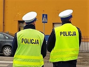 w rejonie przejścia dla pieszych stoją dwaj policjanci, na głowach mają czapki Wydziału Ruchu Drogowego, a na sobie odblaskowe kamizelki