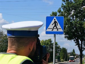 policjant stoi przy przejściu dla pieszych, urządzeniem do pomiaru prędkości mierzy prędkość, z jaką poruszają się pojazdy po drodze