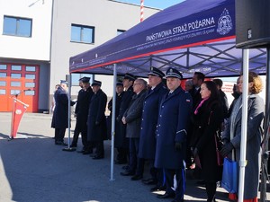 Na placu przed komendą straży pożarnej, pod specjalnym namiotem stoją zaproszeni na uroczystość goście, część ma na sobie mundury galowe policyjne, a także strażackie. Obok funkcjonariuszy stoją inne osoby, przedstawiciele władz miasta, powiatu, komendant wojewódzki PSP i inni zaproszeni na uroczystość goście.