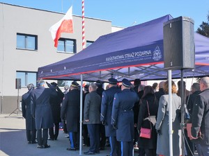 zaproszeni na uroczystości goście stoją frontem do flagi państwowej. Funkcjonariusze oddają honor.