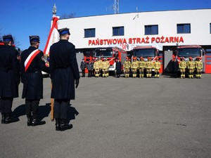 poczet sztandarowy stoi tyłem, na przeciwko nich stoją zaparkowane wozy strażackie, przed nimi na baczność stoją strażacy