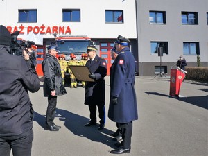 podczas uroczystości przed budynkiem PSP stoją strażacy w mundurach, na przeciwko nich stoją komendanci i władze samorządowe. mężczyzna stoi tyłem i kameruje strażaków.