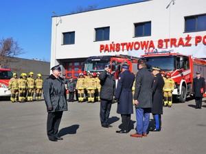 przed budynkiem straży pożarnej trwa uroczystość, strażacy w szykach stoją przed zaparkowanymi wozami strażackimi, na środku stoją dwaj strażacy, przy drugim stoi komendant PSP i komendant wojewódzki PSP, a także przedstawiciele władz samorządowych. Strażak oddaje honor.