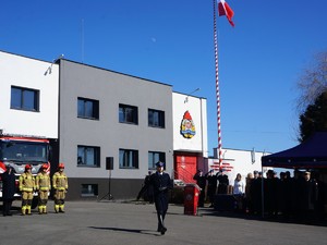 na placu trwa apel, część gości stoi pod namiotem, strażacy stoją w szyku przed zaparkowanymi wozami strażackimi, strażak w galowym mundurze maszeruje.