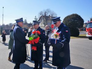 Komendant wodzisławskiej Policji i jego zastępca składają życzenia  komendantowi PSP. Komendant w rękach trzyma bukiet kwiatów, jego zastępca trzyma puchar pamiątkowy i teczkę z logo wodzisławskiej komendy. Mężczyźni mają na sobie mundury galowe.