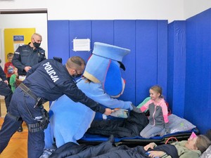 dziewczynka siedzi na łóżku, łapkę do niej wyciąga pies Sznupek, a policjant przekazuje kolorowankę