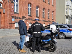 policjant stoi przy policyjnym motocyklu, obok niego stoi młody mężczyzna, trzyma w ręku kartkę papieru, obok stoi zaparkowany radiowóz, w tle budynek Urzędu Miasta Wodzisław Śląski